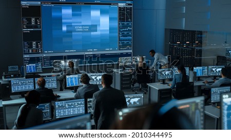 Group of People in Mission Control Center Trying to Establish Video Connection on a Big Screen with an Astronaut on Board of a Space Station. Flight Control Scientists Sit in Front Computer Displays. Royalty-Free Stock Photo #2010344495