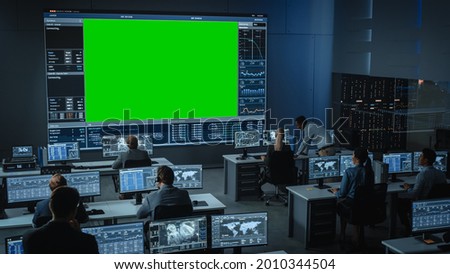 Big Green Screen Horizontal Mock Up in a Mission Control Center Room with Flight Director and Other Controllers Working on Computers. Team of Engineers Work in Monitoring Room Full of Displays. Royalty-Free Stock Photo #2010344504