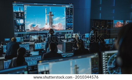 Group of People in Mission Control Center Witness Successful Space Rocket Launch. Flight Control Employees Sit in Front Computer Displays and Monitor the Crewed Mission. Team Stand Up and Watch. Royalty-Free Stock Photo #2010344576