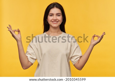Smiling beautiful brunette young woman in white tshirt over yellow keeps hands in mudra zen gesture meditating and doing yoga background