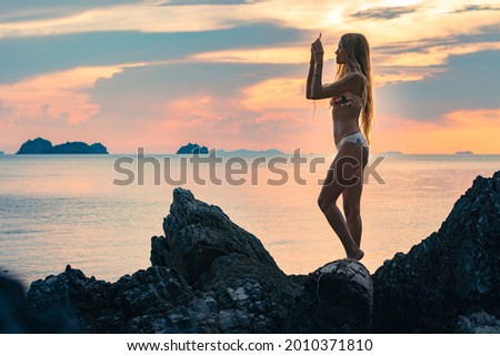 Young happy blonde woman with long hair in a swimsuit stands on a wild beach between cliffs with a smartphone in her hand. Colorful  sunset over sea on the background.