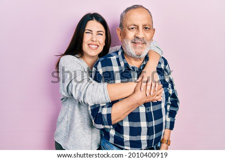 Young brunette woman and senior man standing over pink background. Daughter and father hugging and bonding together as happy family Royalty-Free Stock Photo #2010400919