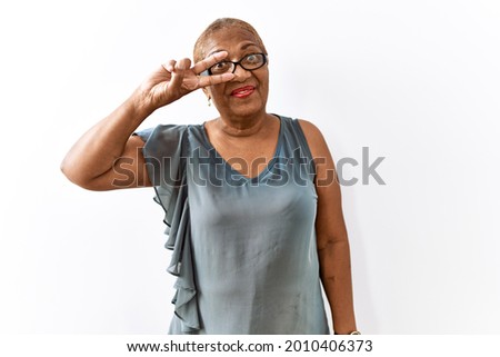 Mature hispanic woman wearing glasses standing over isolated background doing peace symbol with fingers over face, smiling cheerful showing victory 