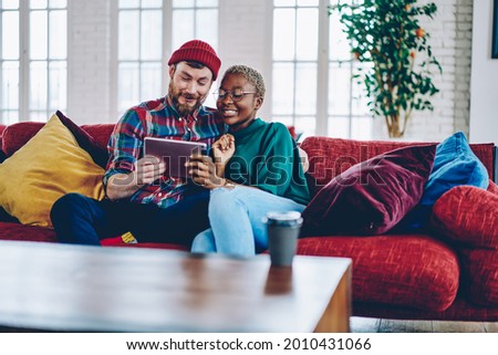 Happy Caucaisan boyfriend and African American girlfriend in glasses enjoying online shopping on website using wireless internet on touch pad, multiracial hipsters watching funny video during weekend