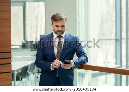 Serious mature businessman in suit standing at office hall and typing a message on his mobile phone