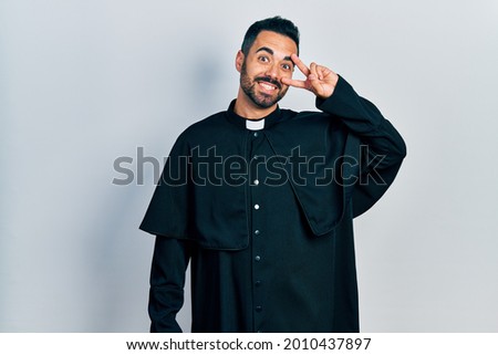 Handsome hispanic man with beard wearing catholic priest robe doing peace symbol with fingers over face, smiling cheerful showing victory 