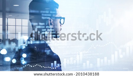 Over the shoulder shot of businessman in glasses and suit looking at financial graphs, candlestick, analytics for investment solution. Double exposure. Modern panoramic office interior on background