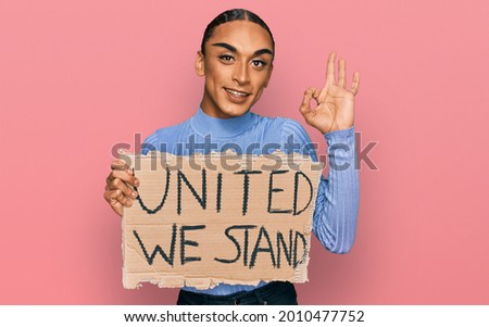Hispanic transgender man wearing make up and long hair holding united we stand banner doing ok sign with fingers, smiling friendly gesturing excellent symbol 