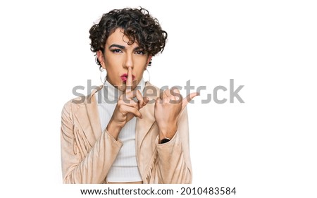 Handsome man wearing make up and woman clothes asking to be quiet with finger on lips pointing with hand to the side. silence and secret concept. 