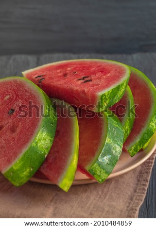 The photo shows a picture of fresh and juicy food. These are watermelon slices with dark seeds. Slices of watermelon are lying on a wooden rustic table with a brown towel