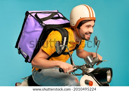 Young courier, delivery man in uniform with thermo backpack on a moped isolated on blue background. Fast transport express home delivery. Online order. Royalty-Free Stock Photo #2010495224