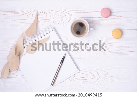 Composition from a blank diary with a spring, a cup of coffee and macaroons and beige dried leaves on a white shabby wooden table. Cozy flat layout.
