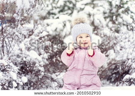 cute caucasian little girl in warm clothes showing thumb up in snowy winter weather