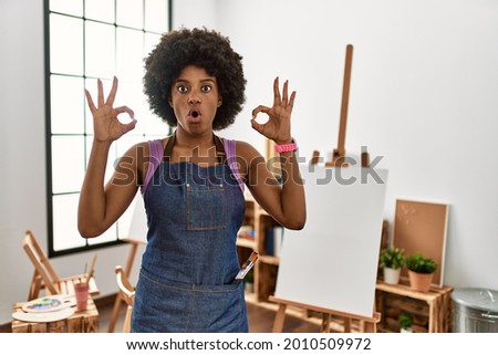 Young african american woman with afro hair at art studio looking surprised and shocked doing ok approval symbol with fingers. crazy expression 