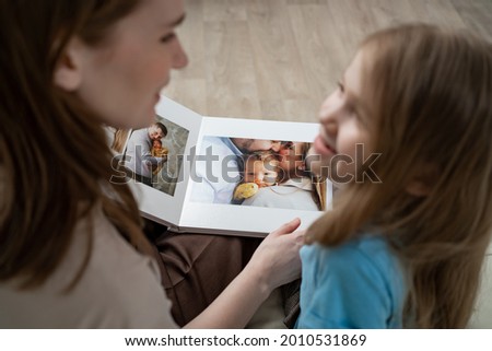 mother and daughter at home on the couch leafing through and looking a book with photos from a family photo shoot with a newborn baby. memory of important moments of life in the photo album. Royalty-Free Stock Photo #2010531869