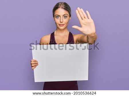 Beautiful blonde woman wearing sportswear holding white banner with open hand doing stop sign with serious and confident expression, defense gesture 