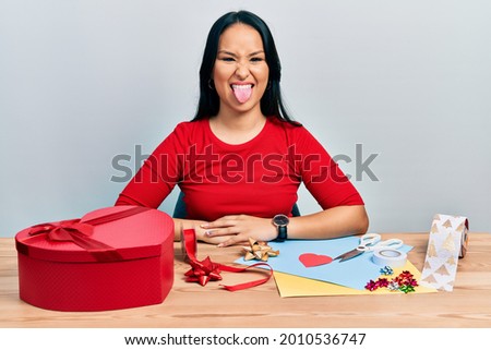Beautiful hispanic woman with nose piercing doing handcraft creative decoration sticking tongue out happy with funny expression. emotion concept. 