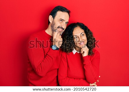 Middle age couple of hispanic woman and man hugging and standing together with hand on chin thinking about question, pensive expression. smiling and thoughtful face. doubt concept. 