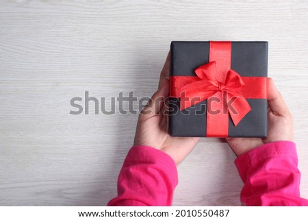 Hands holding black giftbox with red bow on wooden background. Flat lay style. Copy space for text