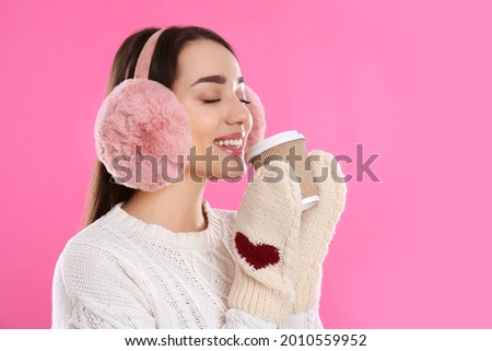 Beautiful young woman in earmuffs with cup of drink on pink background Royalty-Free Stock Photo #2010559952