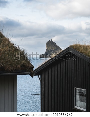 A vertical photo of a rock erupting through the water through the prism of houses in Bour, Faroe Islands