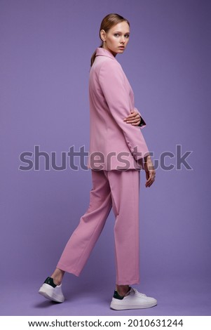 Fashion photo of a beautiful elegant young woman in a pretty soft pink oversized suit, jacket, pants, sneakers posing over purple, lavender, lilac background. Studio Shot.