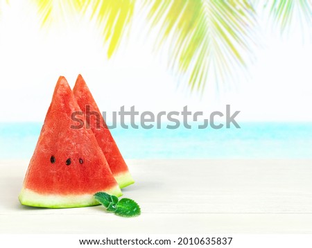 Two triangular slices of watermelon and mint on table top on blurred background of blue sea beach and palm tree at hot sunny day. Stand counter for product display or visual layout design, copy space.