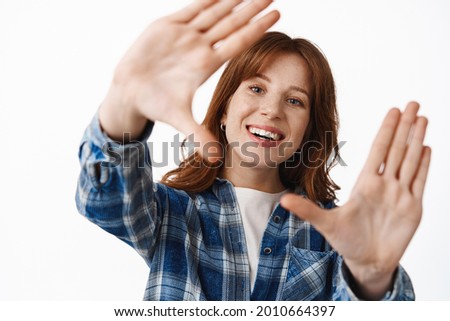 Close up of smiling redhead girl with freckles and white smile, look through hand frames gesture, taking shot of moment, searching for perfect angle, daydreaming, standing over white background