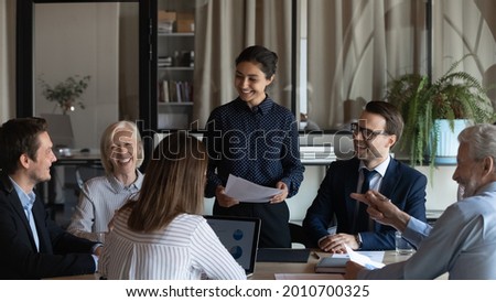 Happy diverse team of different ages with Indian business leader meeting in office, talking, discussing project, brainstorming, laughing. Female coach and employees having fun on training Royalty-Free Stock Photo #2010700325