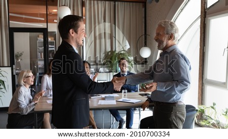 Happy older male team leader giving handshake to excited employee, welcoming new team member, congratulating on good work, hiring for job, career promotion. Team clapping hands and laughing Royalty-Free Stock Photo #2010700334