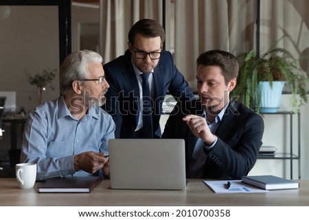 Male business team working on project together, meeting and talking at computer. Office employees of different generations watching and discussing content on laptop, sharing workplace for teamwork Royalty-Free Stock Photo #2010700358