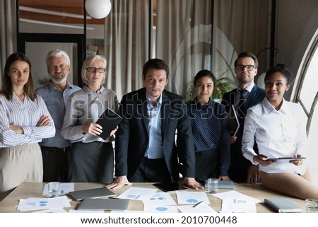 Confident diverse employees of different age standing at meeting table together, looking at camera. Multiethnic young and senior interns, students, mentor, teacher team portrait. Corporate head shot Royalty-Free Stock Photo #2010700448