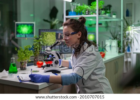 Biologist scientist looking at leaf sample using medical microscope while writing gmo expertise on notepad. Chemist examining biological discovery on plant working in pharmaceutical laboratory.