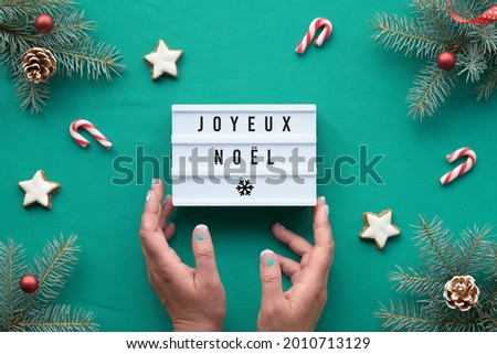 Light box with text Joyeux Noel - Merry Christmas in French language. Hands hold the box. Xmas background, top view on fir twigs with red decorations on vibrant turquoise textile tablecloth.