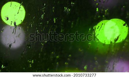 Drops of night rain on dirty glass window with green bokeh lights of city traffic. Rainy weather urban background. Intentional motion blur lights