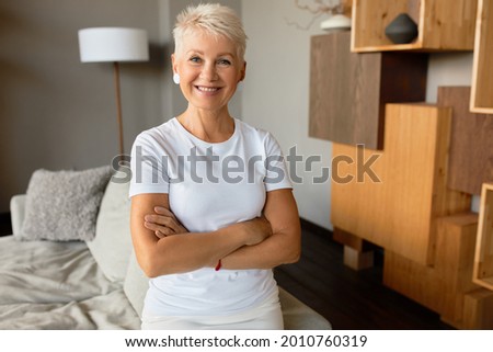 Friendly mature woman with pleasant smile meet guests in living room of countryhouse. House with scandinavian wooden interior for rent on holiday. Welcome, housing rental, vacation concept Royalty-Free Stock Photo #2010760319