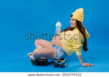 a pregnant girl in yellow clothes with a glass of juice sits on a skateboard on a blue background.