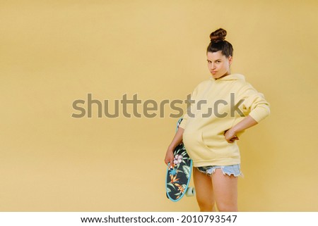 Pregnant girl in a yellow jacket with a skateboard in her hands on a yellow background.