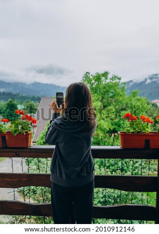 Brunette girl in a sports suit on the balcony takes pictures by phone on the mountain. Girl with a phone on a wooden balcony with red flowers.