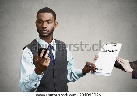 Closeup portrait serious businessman signing contract without looking at document, keeps reading news on smart phone holding mobile isolated grey background. Human face expression corporate executive 