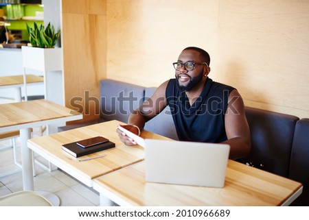 Happy dark skinned male blogger with touch pad and laptop technology spending weekend leisure in cafe interior enjoying time for web networking, smiling African American IT professional in glasses