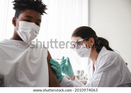 A female doctor or nurse wearing mask,glove and face shield is injecting the coronavirus 19 vaccine on the shoulder of African-American man to immunize. Concept of preventing the spread of COVID-19.