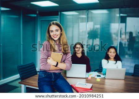 Half length portrait of happy female employee holding education copybook in hands and smiling at camera while sitting at table desktop with blurred colleagues on background, woman with textbook posing