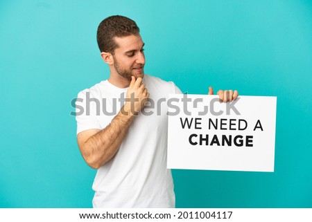 Handsome blonde man over isolated blue background holding a placard with text We Need a Change and thinking