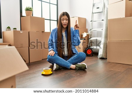 Young chinese girl sitting on the floor at new home pointing finger up with successful idea. exited and happy. number one. 
