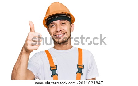 Hispanic young man wearing handyman uniform and safety hardhat smiling happy and positive, thumb up doing excellent and approval sign 