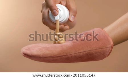 Dosing bronzing lotion or tanning cream from a flask with a doser to a pink tan applicator glove. High quality close-up studio photo image beige background. Royalty-Free Stock Photo #2011055147
