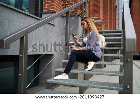 Casual dressed female tourist using cellular application for reading travel publication on website resting at urban stairs in city, Caucasian student with coffee to go connecting to 4g internet