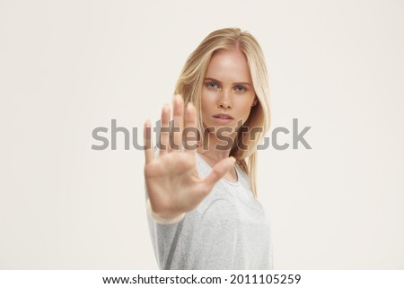 Young serious european girl with stop gesture. Beautiful blonde female teenager with blue eyes wear white t-shirt and looking at camera. Isolated on white background. Studio shoot