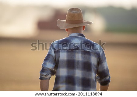 Attractive young farmer wearing straw hat and plaid shirt. Male agronomist with back to camera walking in field. during harvest Royalty-Free Stock Photo #2011118894
